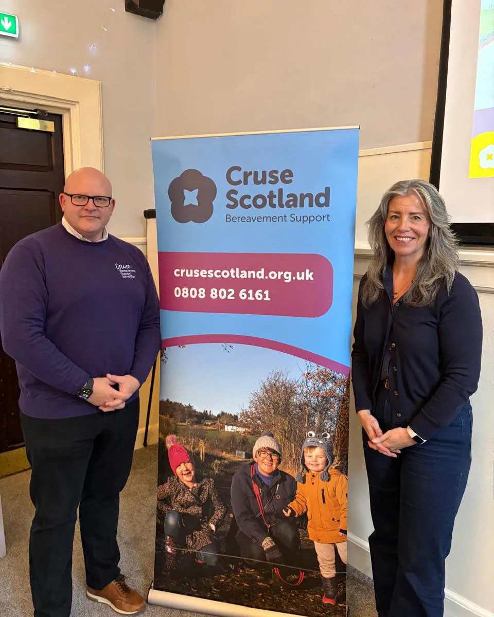 Cruse Isle of Man chair John and Cruse Scotland chair Elena Fry are standing on either side of a Cruse Scotland banner in the recoption area of a hotel/function room.
