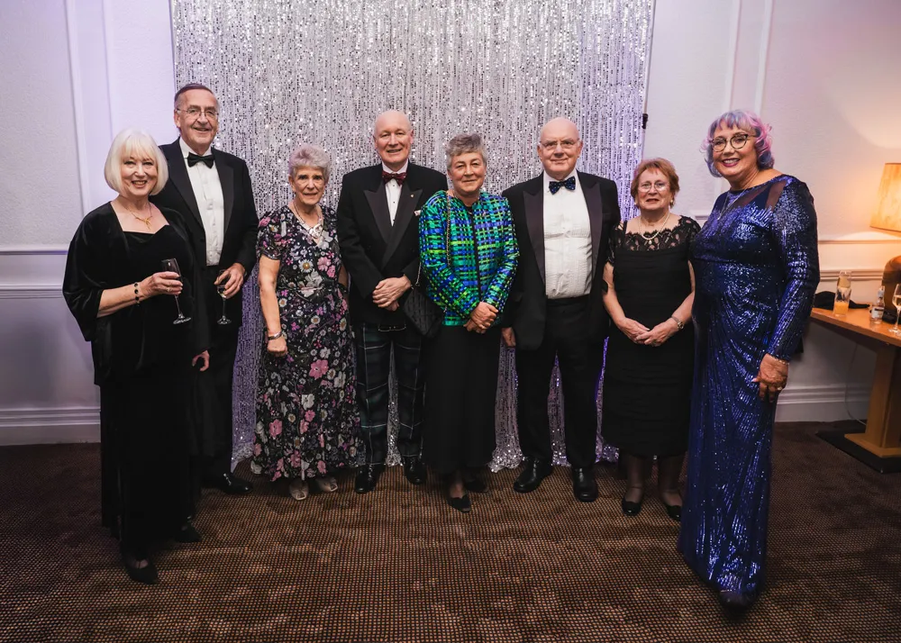 Eight guests at the dinner are dressed in evening wear and are standing in front of a sparkly background on the wall. All are smiling for the camera.