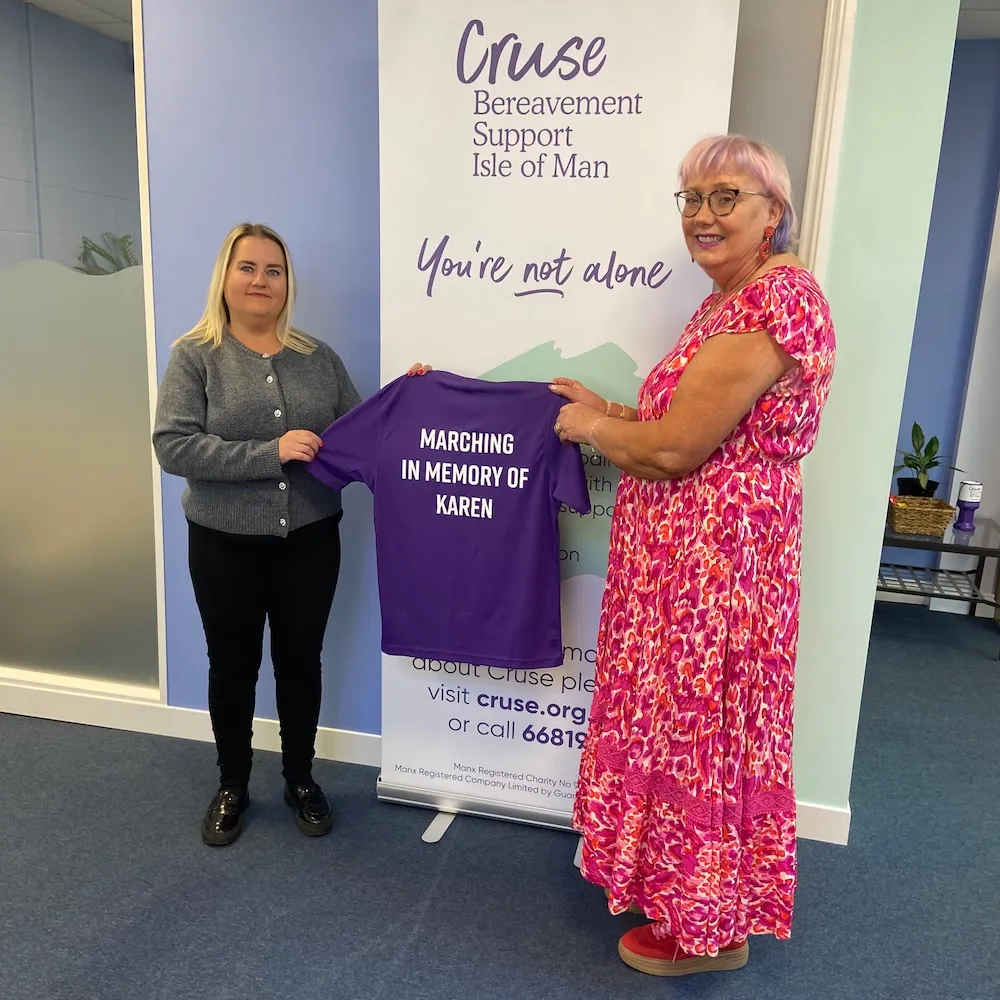 Emma and Mary are standing in front of a Cruse banner and smiling for the camera. Each of them is holding the arm of a purple t-shirt with white writing on it that says 'Marching in memory of Karen'.