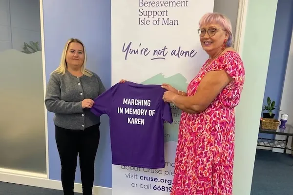 Emma and Mary standing in front of a Cruse banner. Each of them is holding a sleeve of a purple t-shirt which hangs betweeen them. The printed message on the t-shirt says 'Marching in memory of Karen in white letters.