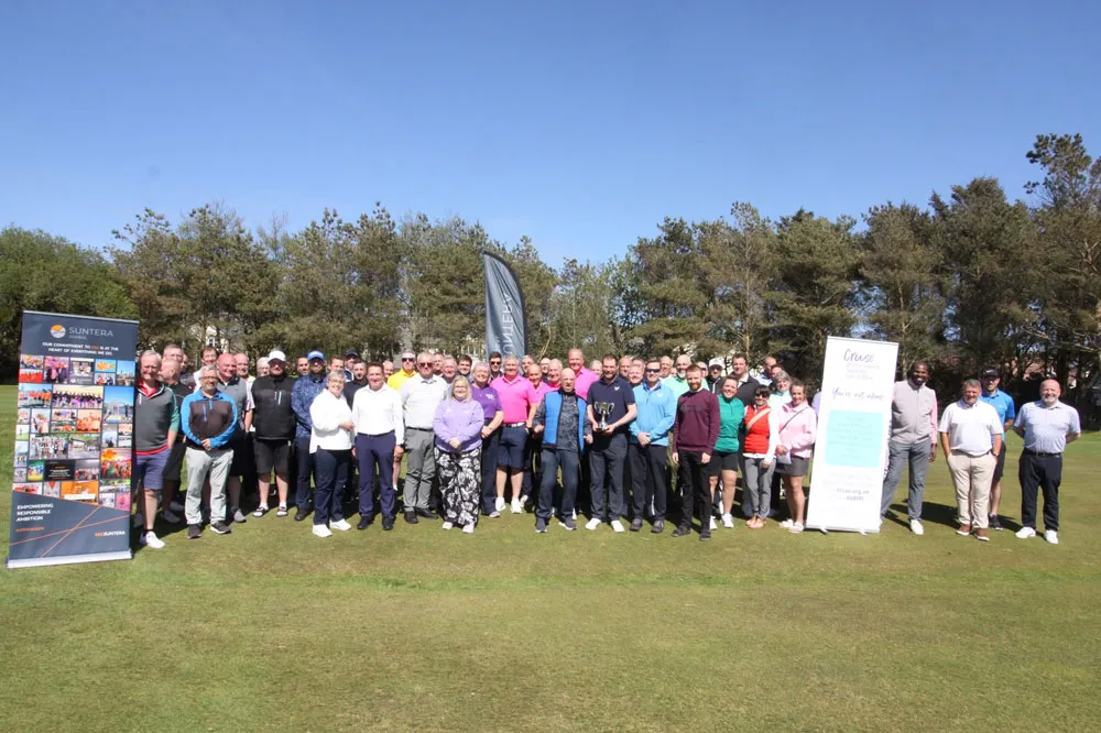 All of the players at the golf day (about 80 people) are standing together in a group shot in front of the 18th green with Suntera and Cruse banners on either side. All are smiling for the camera, and there are trees behind them and a blue sky above them