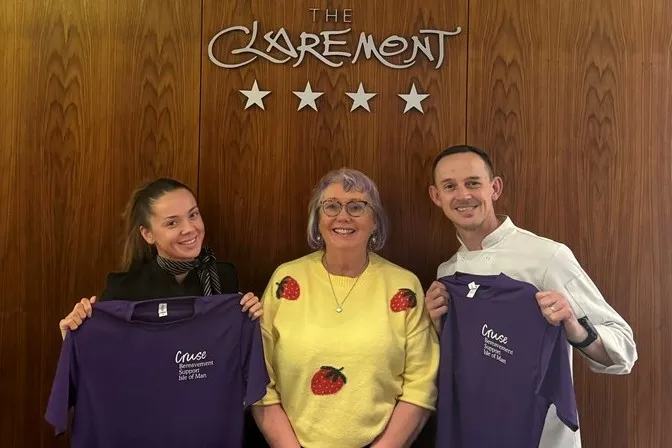 Jade and Les standing with Mary in front of the Claremont sign behind the reception desk at the Claremont Hotel, where they both work. Jade and Les are holding up purple Cruse t-shirts and everyone is smiling for the camera.