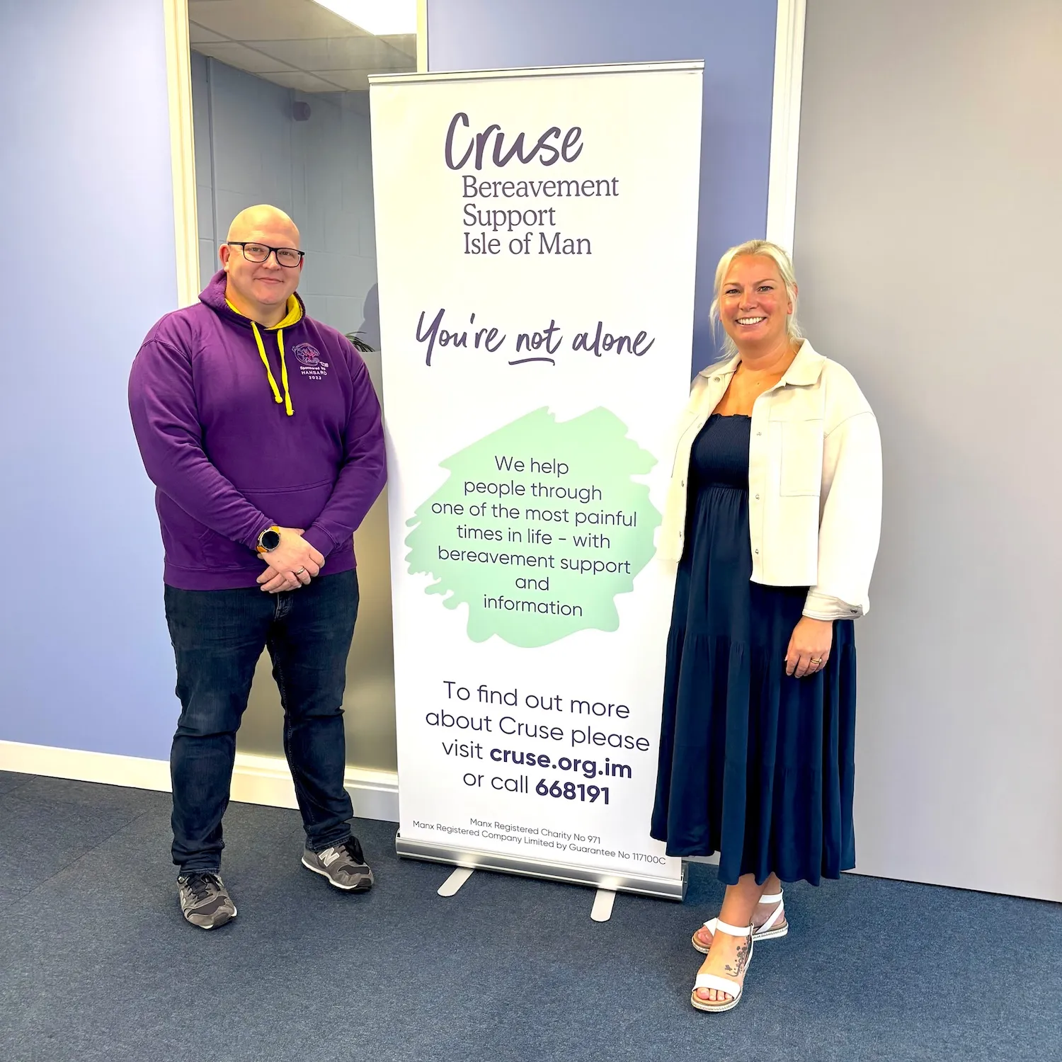 John and Sally are standing either side of a Cruse banner in the Cruse office. Both are smiling for the camera.
