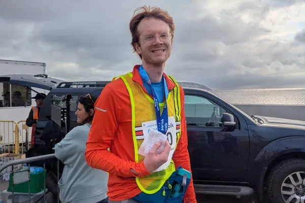 Samuel is standing on Douglas Promenade after finishing the Parish Walk. He;s wearing a bright orange top and a yellow high-vis vest, with a water bottle clipped to his waist. He's holding his timing sheet and his medal is around his neck as he smile for the camera.