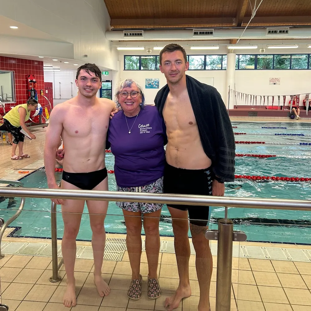 Our chief executive Mary is standign between Joel and Sean. Mary is in a purple Cruse t-short and Joel and Sean are in swimming trunks. All are smiling for the camera with a swimming pool behind them with swimmers in the water.