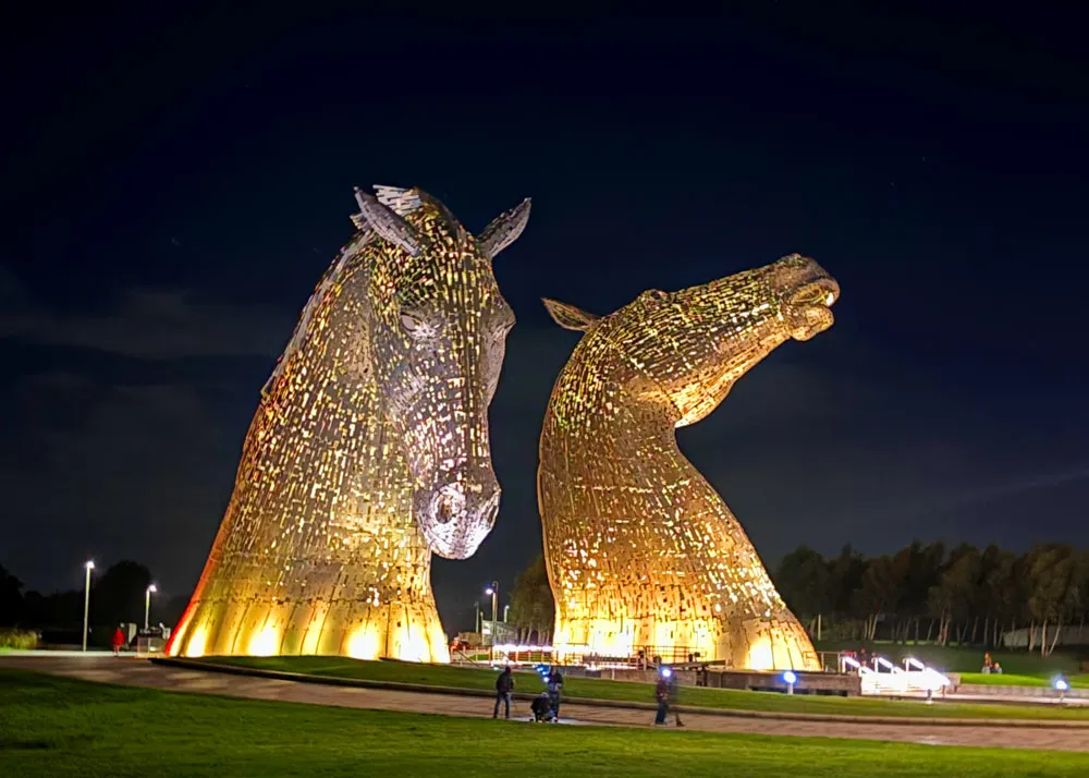 A photo of the Kelpies at night - two 100 ft tall steel sculptures of horses' heads lit up in white lights.